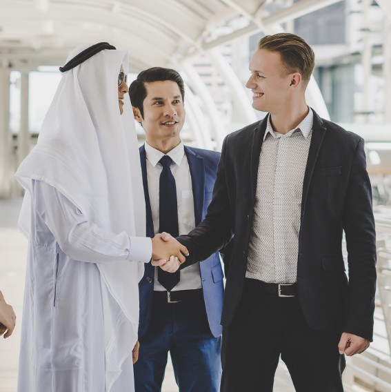 Tres hombres de negocios en un entorno moderno al aire libre, dos de ellos estrechando la mano en señal de acuerdo. Uno de los hombres viste una túnica tradicional árabe, mientras que los otros dos llevan trajes formales.