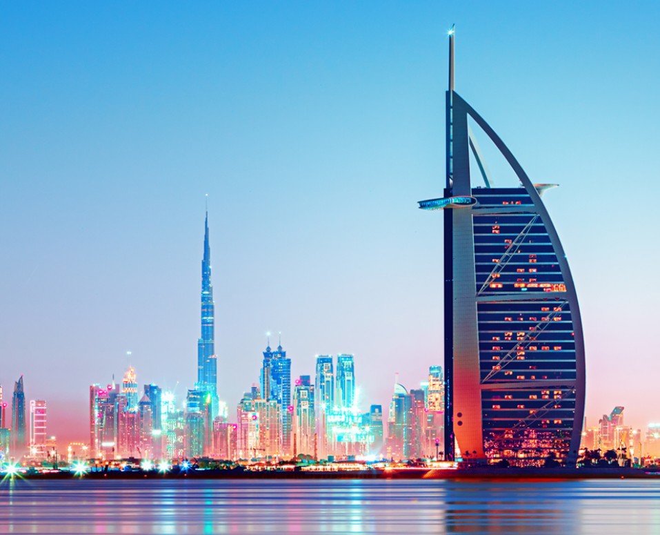 Vista panorámica de Dubái al atardecer, con el icónico hotel Burj Al Arab en primer plano y el Burj Khalifa junto al moderno skyline iluminado en el fondo, reflejándose en el agua.