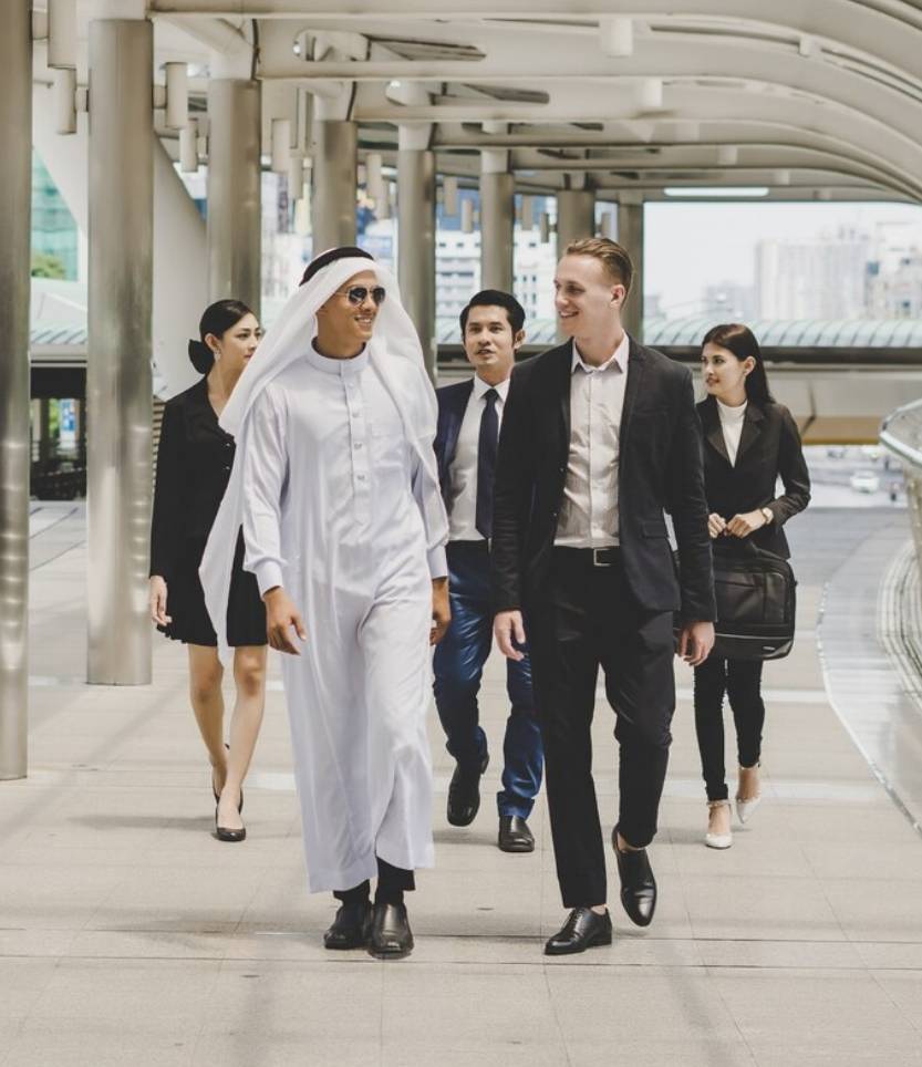 Un grupo de cinco personas camina por un moderno pasillo al aire libre en un entorno urbano. El hombre en el centro viste una túnica blanca tradicional y gafas de sol, mientras conversa con otro hombre de traje negro y camisa blanca. Detrás de ellos, tres personas con vestimenta formal, dos mujeres y un hombre, los siguen. El fondo muestra edificios y una estructura con techo curvo y pilares metálicos.